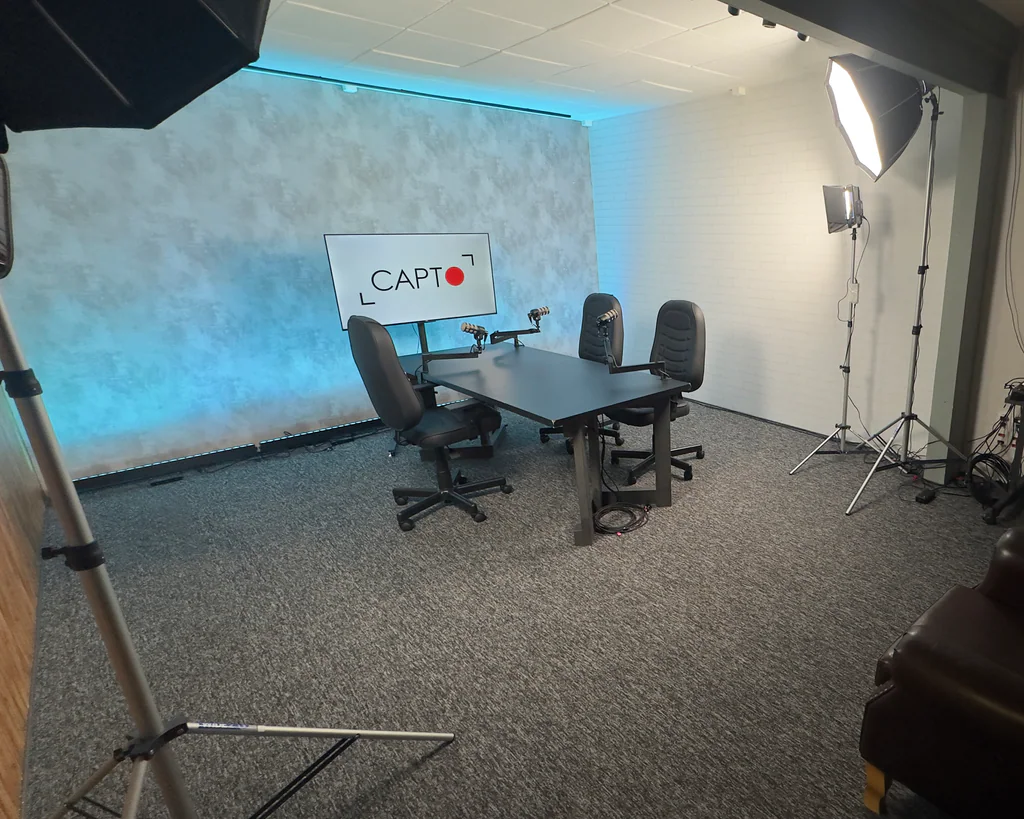 Estúdio de podcast da Capto com mesa, três cadeiras, microfones, TV com logo e softboxes de iluminação