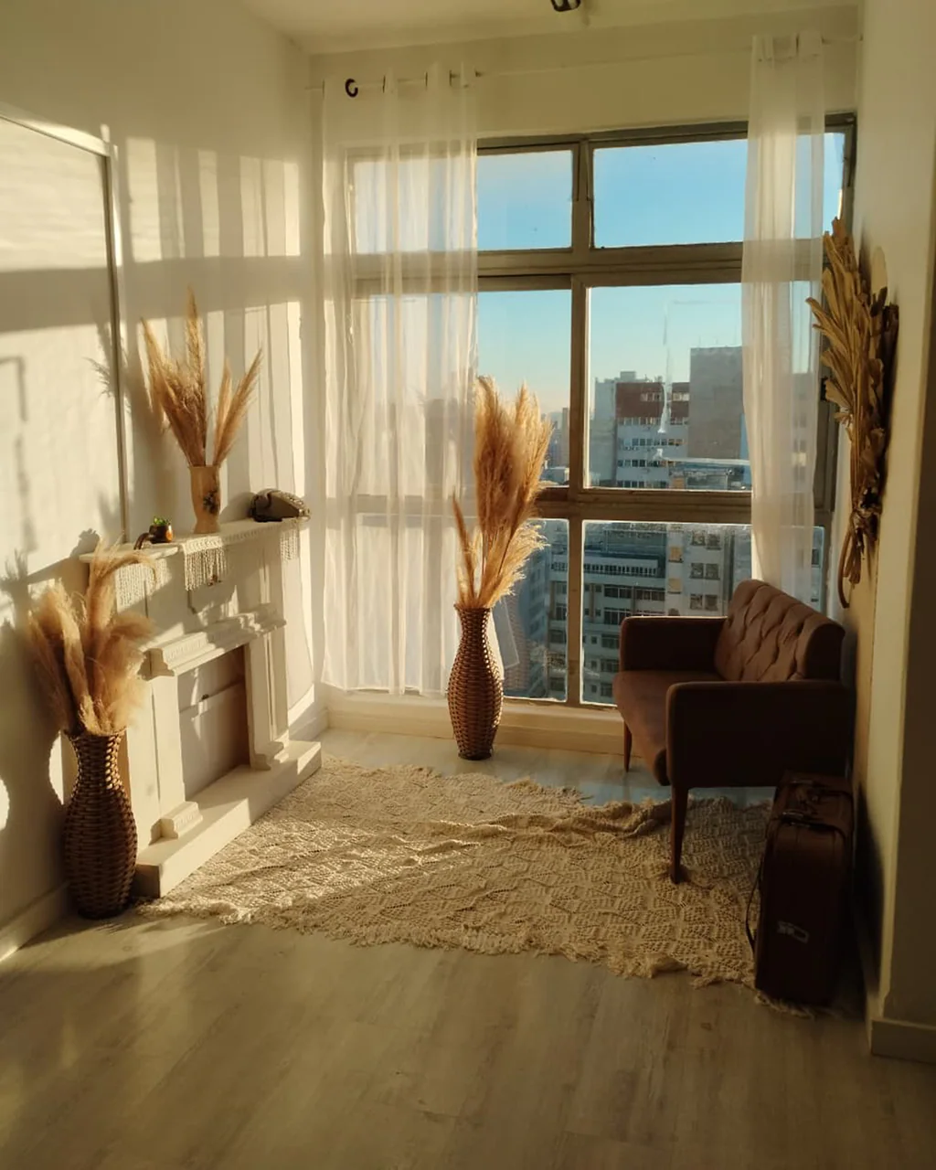 Ambiente com lareira, vasos de capim-dos-pampas, poltrona marrom e vista da cidade ao entardecer no Estúdio Grimm