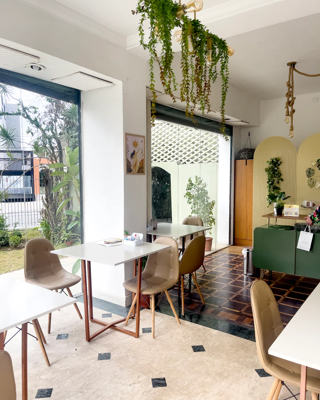 Área do café com mesas brancas, plantas suspensas e balcão verde no Espaço Iluminar