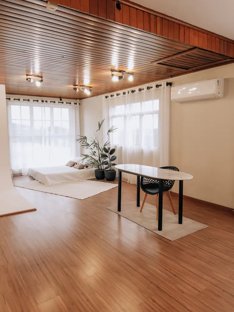 Ambiente amplo do Macchiato com cama junto à janela, mesa oval com cadeira e teto de madeira ripada