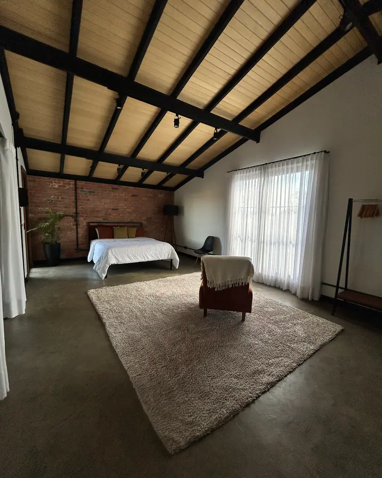Quarto amplo com teto industrial em madeira e ferro, parede de tijolinho, cama e poltrona sobre tapete no Mammü estúdio