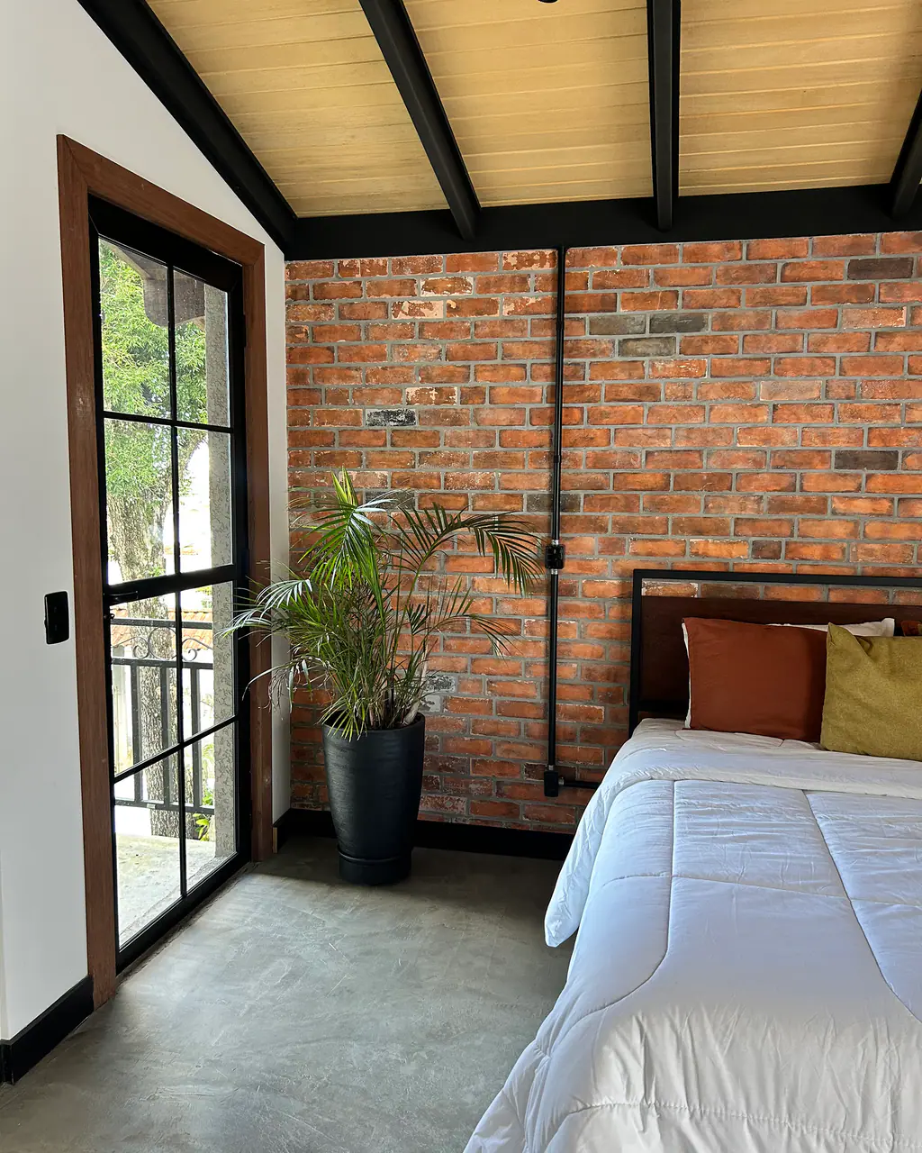 Quarto com parede de tijolo aparente, cama de casal, planta e porta de vidro com vista para a varanda no Mammü estúdio
